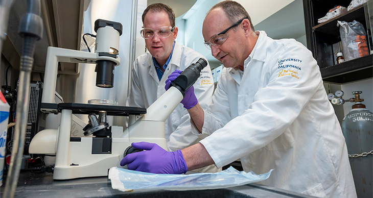 Dominik Haudenschild is wearing a white gown, purple gloves, and a pair of goggles. He is looking through an electronic microscope. Blaine Christiansen is wearing a white gown and goggles and standing next to Haudenschild. 