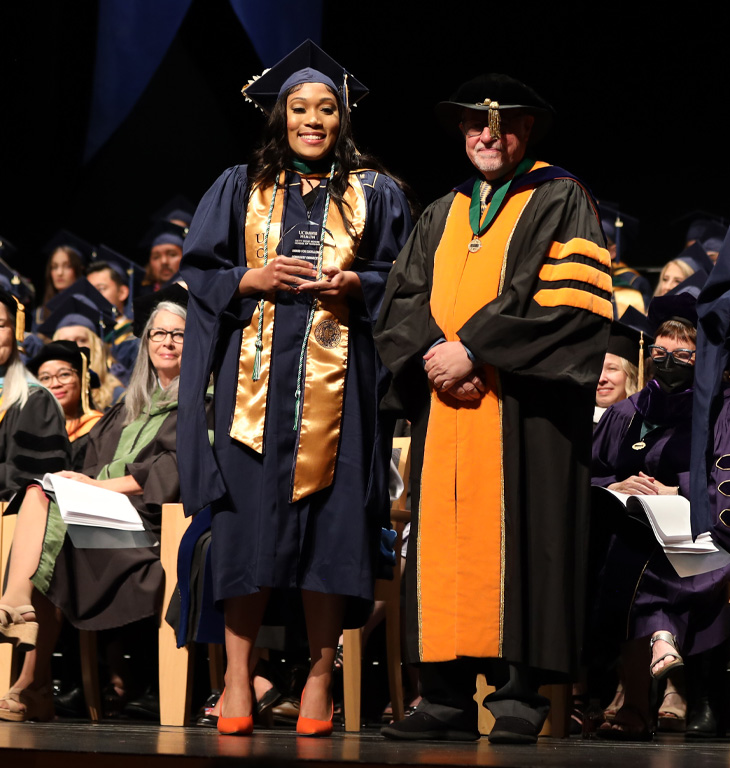 Aliyah Bell, left, stands holding award next to school dean on stage smiling at camera in audience