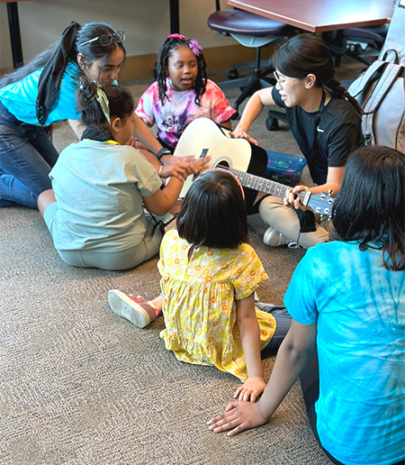 a small group of young people sits around a person who is playing guitar. A young woman holds the hand of one of the young people as they reach for the guitar. 