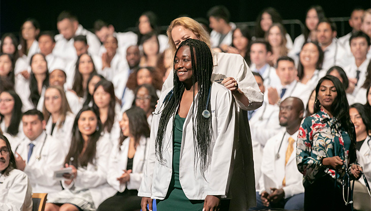 Woman with dark skin standing with stethoscope around her shoulders and another woman standing behind her.