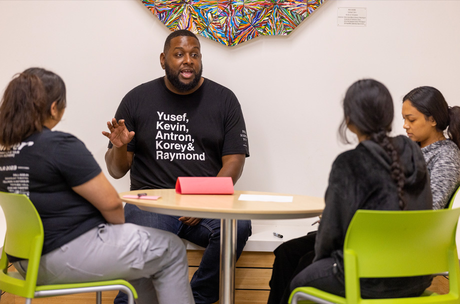 Man with outstretched hand speaks with three students as they sit at a circular table