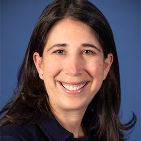 Woman with dark brown hair and brown eyes, wearing a dark blue suit jacket, smiles widely for a portrait.