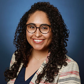 Portrait of woman wearing glasses and with dark, curly hair past her shoulders.