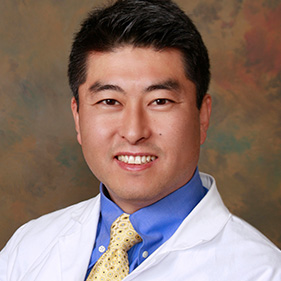 Man with dark hair wearing a white lab coat on gray background