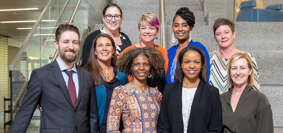 The nine inaugural fellows stand in two rows, shoulder to shoulder on a stairwell, smiling at camera.