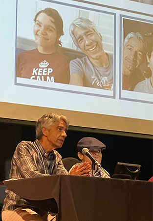 A man and younger man wearing a hat sit on a stage behind a lectern with microphones. Behind them is a PowerPoint presentation that includes images of themselves. 