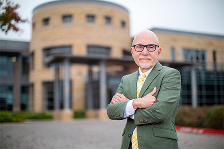 A man wearing a green suit and yellow tie, stands with his arms crossed in front of the UC Davis MIND Institute, a large reddish-brown building.