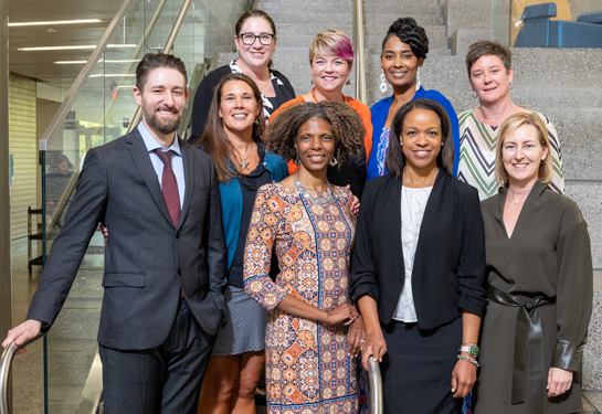 The nine inaugural fellows stand in two rows, shoulder to shoulder on a stairwell, smiling at camera.