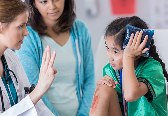 Girl with scraped knee holding blue ice pack to her head as doctor with white coat holds up four fingers and woman stands in the background