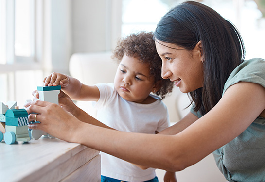 A woman with medium-length dark hair, wearing a green short-sleeved shirt leans next to a young child with curly dark hair, as they play