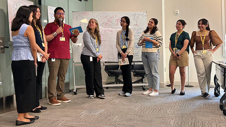 Seven young women and a young man, some holding pamphlets, stand together in a classroom to discuss a class assignment 