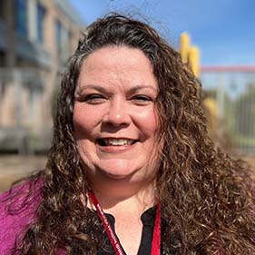 A woman with long curly brown hair wearing a purple sweater and black shirt and smiling stands outside on a playground.