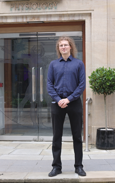 A man wearing a blue shirt and black pants stands in front of a building with the word “PHYSIOLOGY” over an entrance with glass doors.