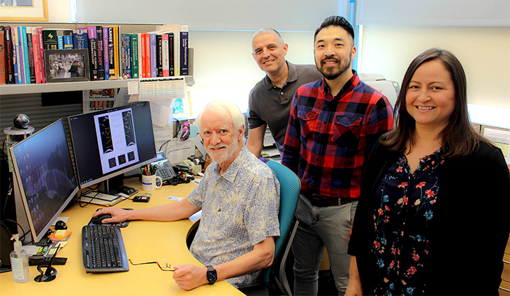 A man sits at a desk in front of a computer monitor with three people standing behind him.