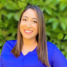 A woman with long dark brown hair, wearing a bright blue shirt stands outside in front of a wall of greenery, smiling. 