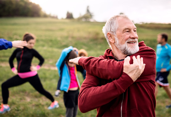 Older man in foreground stretching arm over shoulder while others in exercise clothing stretch in field