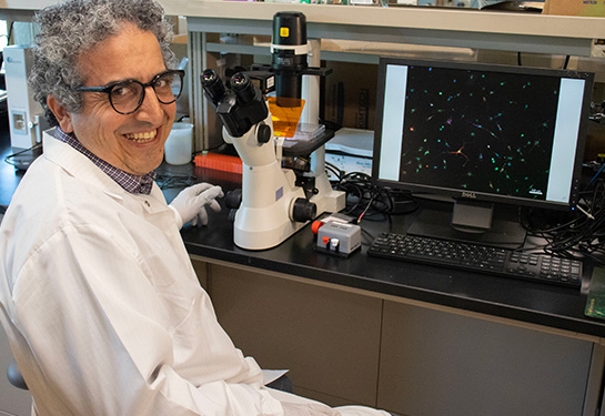 Roy Ben-Shalom wearing white lab coat and gloves, sitting in front of an electronic microscope and a computer in his lab at the MIND Institute