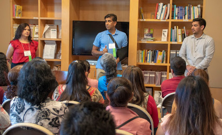 Three adults stand at the front of a large room, giving a presentation to dozens of people sitting in chairs and facing toward them. 