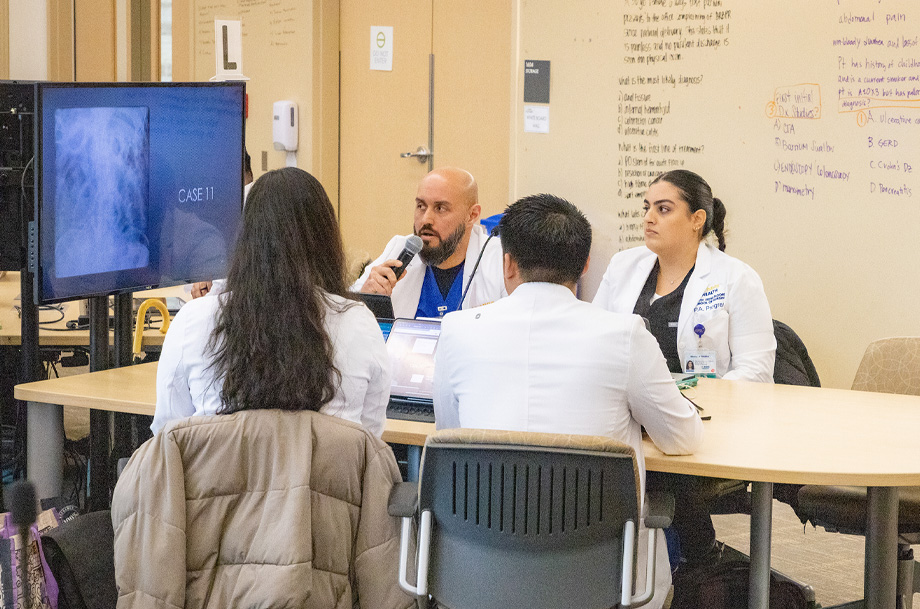 Students in white coats sit at round table looking at X-ray on TV monitor; one student holds microphone