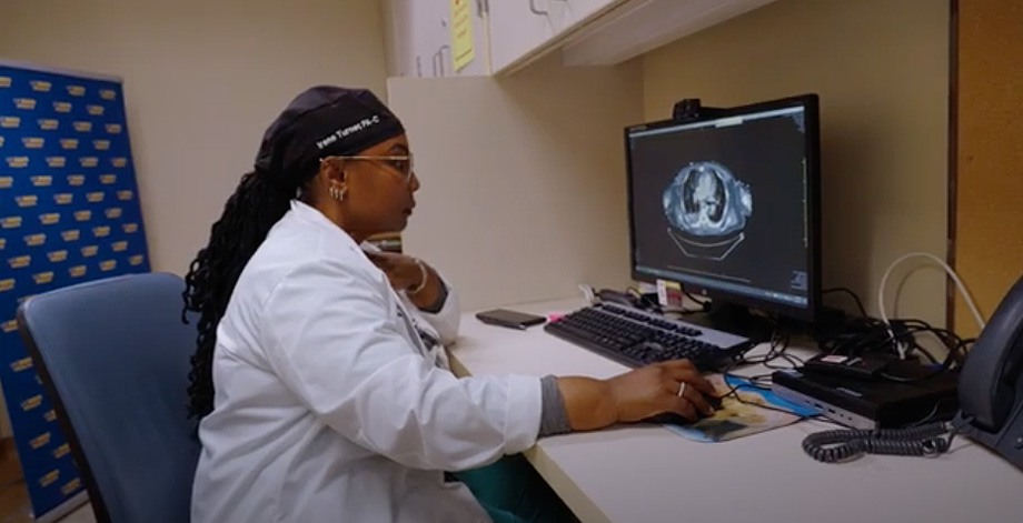 Woman wearing headwrap and white coat sits at computer moving mouse with her hand