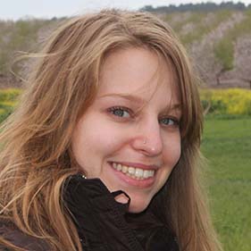 A woman with long brown hair, wearing a brown heavy jacket smiles while standing outside in a field of green with hills in the distance. 