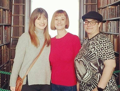 Three women standing together in a library with rows of books