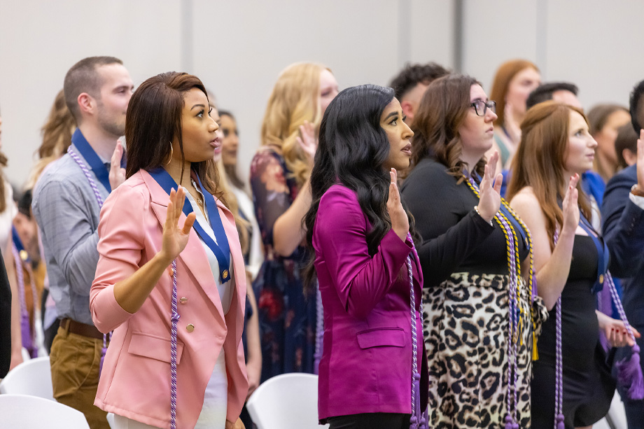 Nursing students stand in rows looking off to right-hand side of frame raising hand and reciting oath