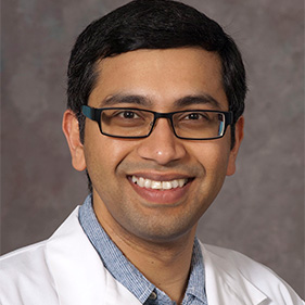 A man with black hair, wearing glasses and a white physician’s coat smiles