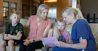 A mother with two young sons sit on a couch in a medical waiting room as a nurse wearing blue scrubs shows them paperwork about vaccinations. 