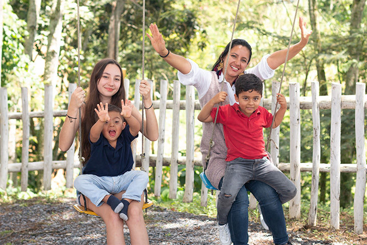 two women sit on swings next to each other; each has a small child on their lap. One child and one woman have their hands in the air. 