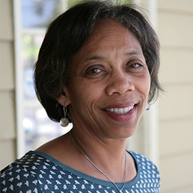 A woman with short dark hair, wearing a blue and white sweater, stands outside near the window of a beige colored building.