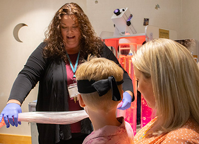 A woman wearing a black long-sleeved cardigan holds a sensory toy made of long plastic strings out to a young boy who’s sitting in his mom’s lap. 