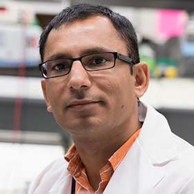 A man with brown hair and glasses wears an orange-striped shirt and a white coat.