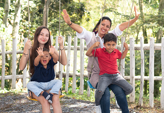 two women sit on swings next to each other; each has a small child on their lap. One child and one woman have their hands in the air.