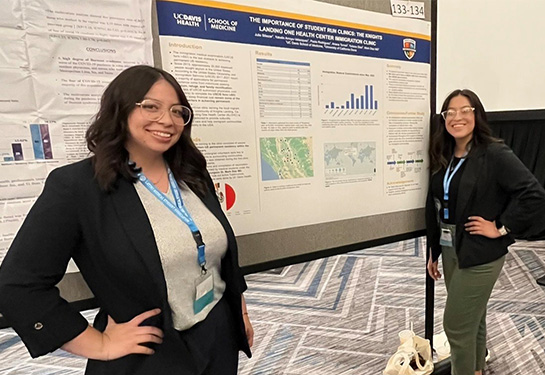 Two young women wearing eyeglasses and dark blazers stand before a white and blue academic poster filled with text and graphics