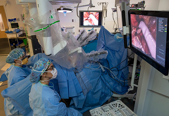 Machine with multiple arms over person laying in bed with two monitors showing surgery