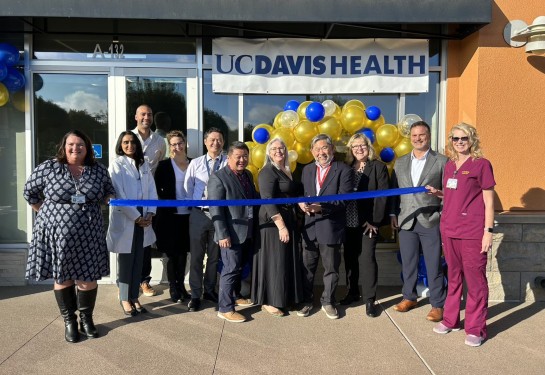 11 people stand outside the front doors of a clinic, with one man holding scissors to cut the blue ribbon in front of them