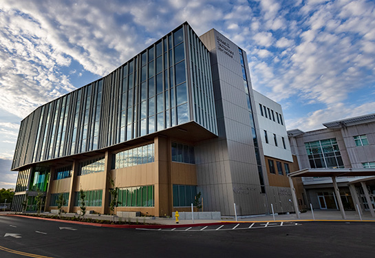 A modern four-story building with reflective glass windows is seen against a blue sky dotted with white clouds. 