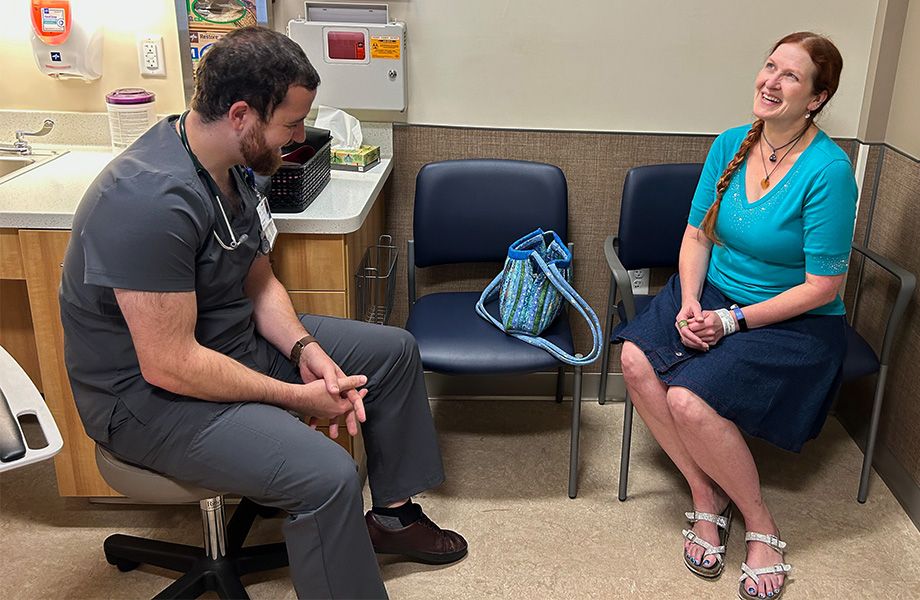 A man with dark hair and beard, in gray scrubs, talks with a woman with long braided hair and blue top in a clinic exam room 