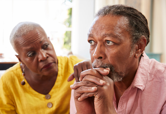  Woman, left, looks at man in a concerned fashion with her hand on his shoulder while he stares off into the distance with hands clasped below his chi