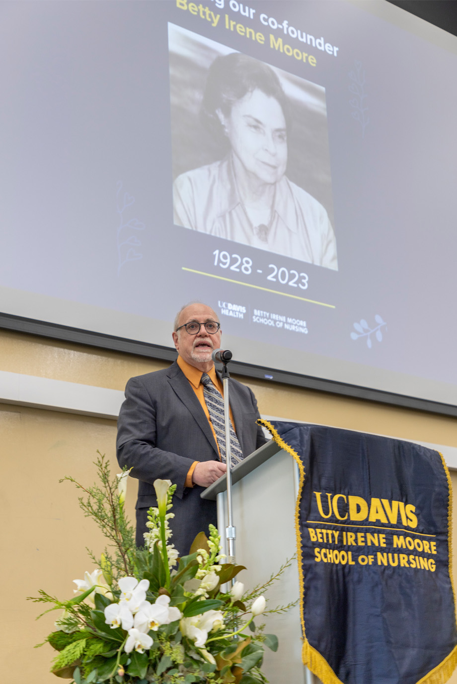 Dean stands at podium speaking into microphone with image of Betty Irene Moore on screen behind him