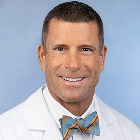 A man with brown hair and glasses, wearing a white doctor’s coat, smiles. 