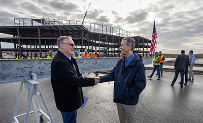 Two men wearing dark coats and pants shake hands in front of a partly constructed building.