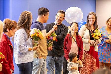 A man in a dark shirt holding a microphone speaks to three people holding colorful bouquets of flowers