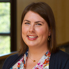 A smiling woman with medium-length brown hair wears a navy sweater and white flowering shirt.