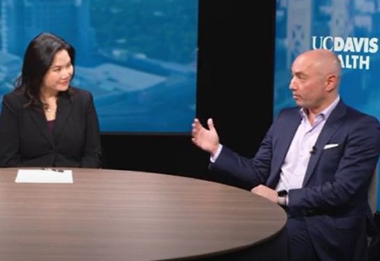 A woman in dark blazer seated at a round table interviews a man in a suit before a blue background and the &#x201c;UC Davis Health&#x201d; logo