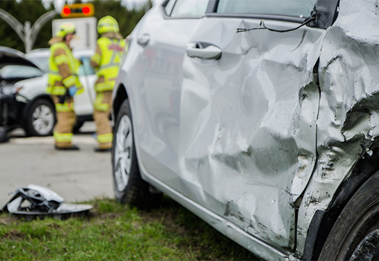 Damaged gray car with three firefighters wearing yellow jackets in the background.