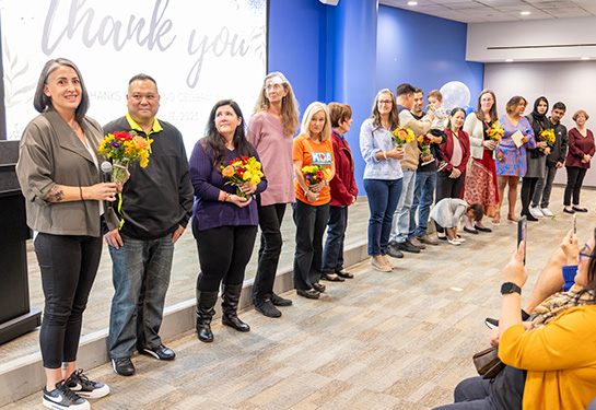 A group of people stand shoulder-to-shoulder forming a long line in a room with a sign stating &#x201c;thank you&#x201d; in the background