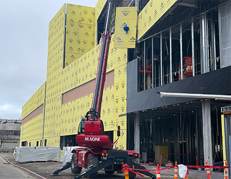 A red crane hoists a yellow wall pane with blue ribbon, watched by people in neon orange vests 