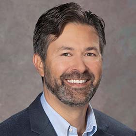 A man with short black hair wearing a dark blue shirt smiles for a portrait